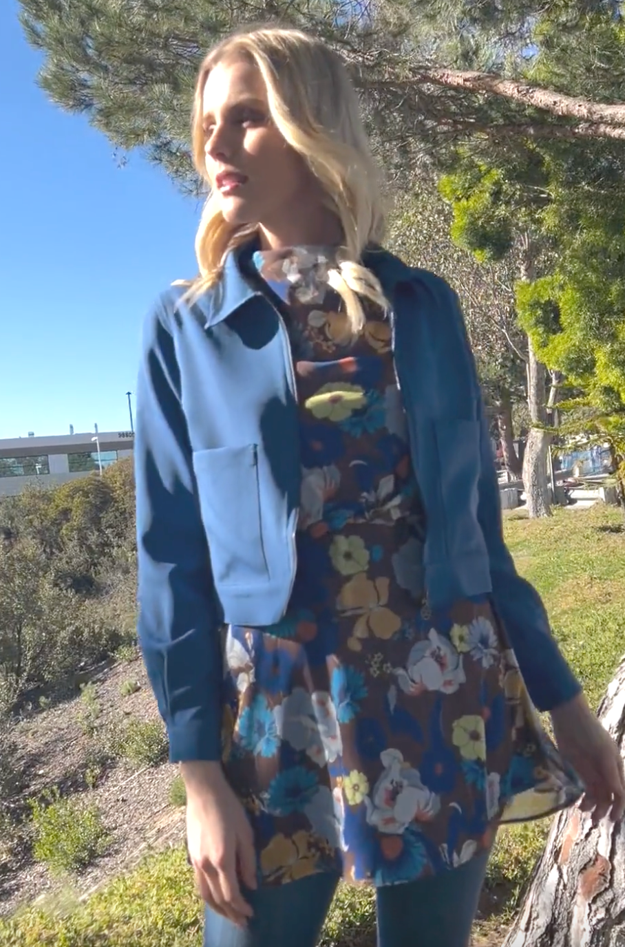 view of woman in park wearing a blue cropped jacket with a floral top at a park. 