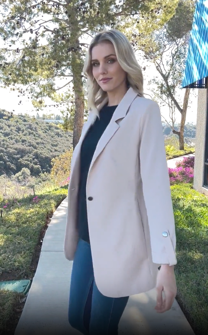 Side view of woman wearing a cream blazer jacket with black top inside at a park..