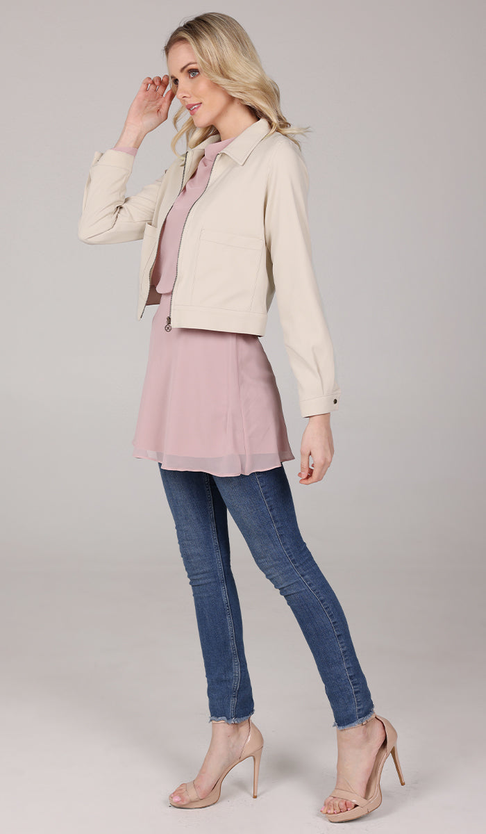 side view of a woman wearing a cream cropped jacket with a blush top and blue jeans.