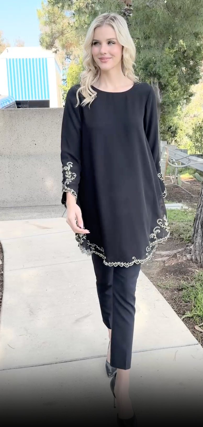 Woman walking outside wearing Meena tunic in Black with black pants and heels. 