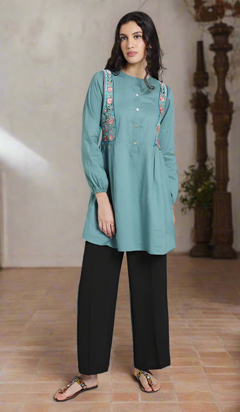 Woman in living room wearing Marzo tunic in Seagreen with black pants and sandals. 