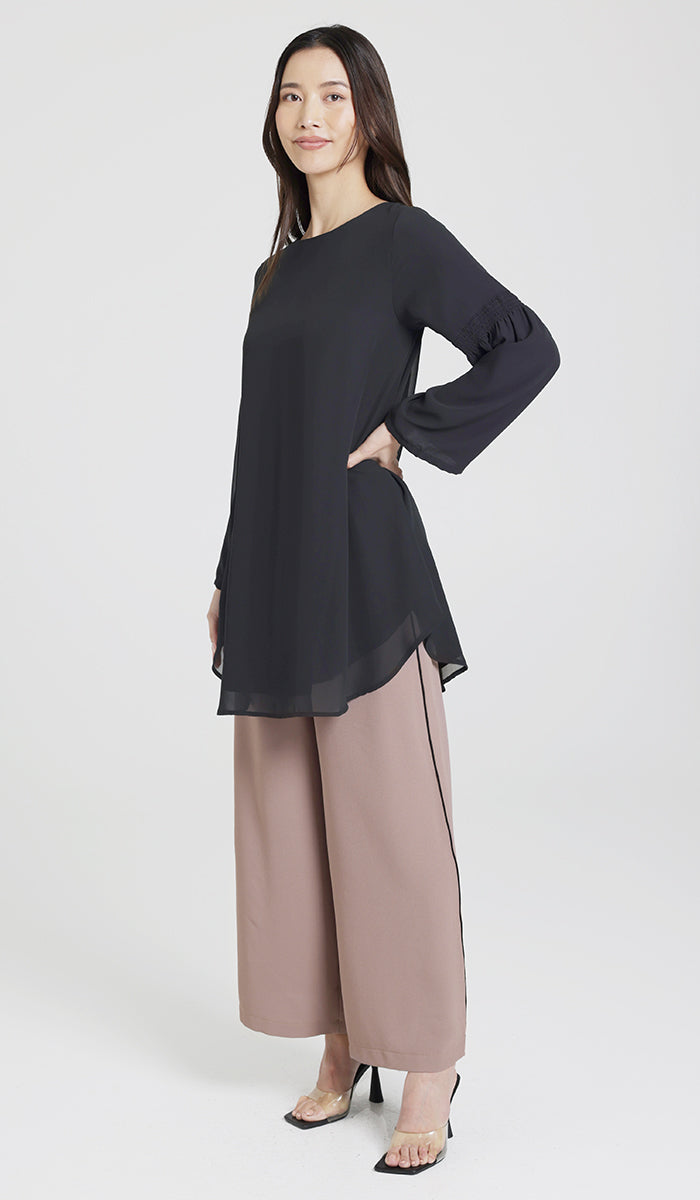 Model smiling posed hand on hip wearing black tunic, brown pants, and black heels.