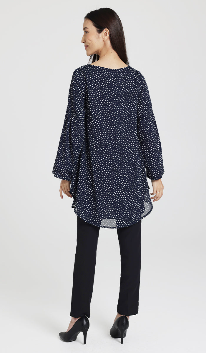 Back view of model wearing  modest navy and white polka dot tunic with black pants and heels.