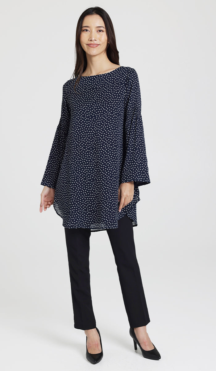 Model posing wearing flowy navy and white dotted tunic with black pants and heels. 