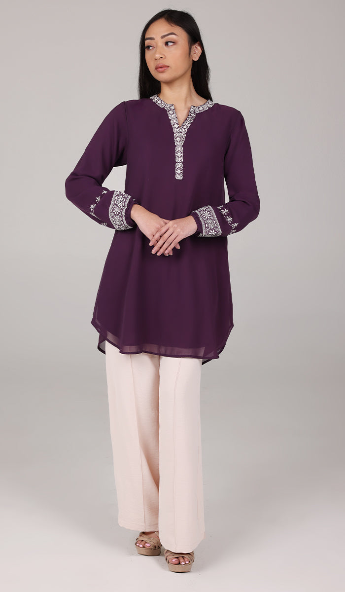 Woman looking to the side wearing Amalie tunic in Eggplant with white pants and heels. 