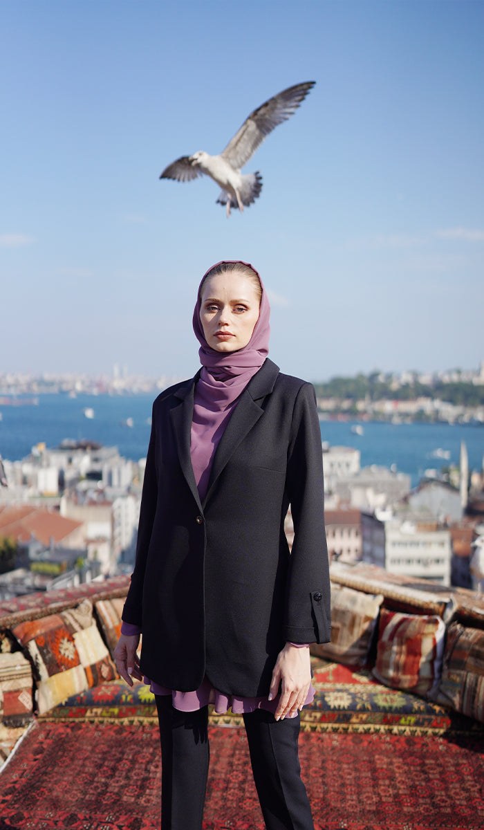 Woman with dusty mauve hijab and top wearing a black blazer with ocean background.