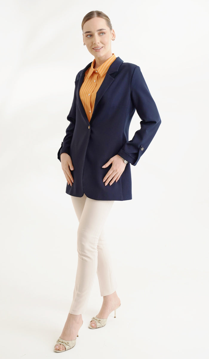 side view of woman wearing an orange top inside a navy blue blazer with cream pants.