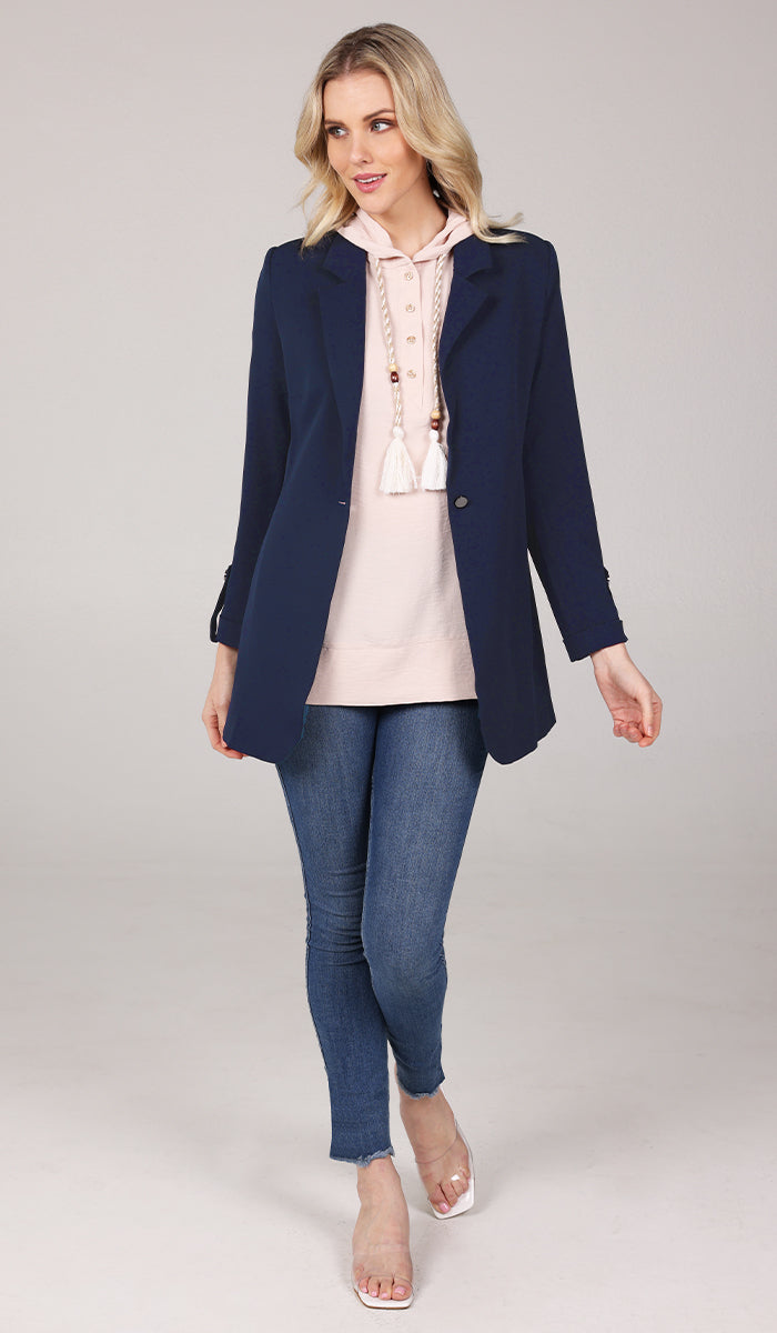 Full body view of woman wearing a cream hoodie inside a navy blue blazer with blue jeans.