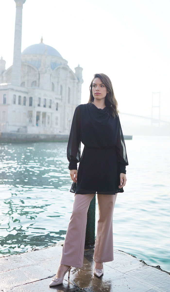 Woman by the water wearing the Myra modest tunic in Black with Lyla pants in Taupe with heels. 